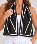 A woman wearing a black cooling towel scarf with white geometric stripes and a white border, styled around her neck over a white tank top and white drawstring pants.
