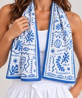 A woman wearing a white tank top and white drawstring shorts with a blue and white patterned cooling towel draped around her neck, photographed against a light neutral background.