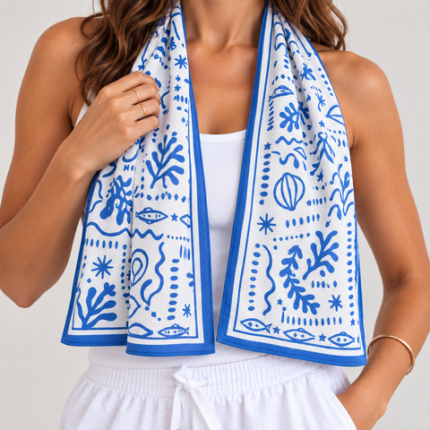 A woman wearing a white tank top and white drawstring shorts with a blue and white patterned cooling towel draped around her neck, photographed against a light neutral background.