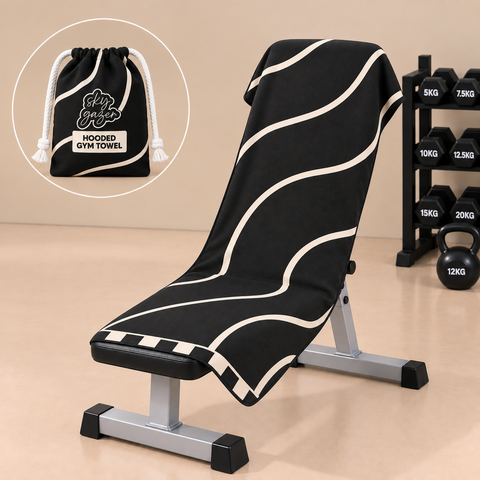 A black hooded gym towel with beige wavy stripes draped over a weight bench, with a circular inset showing the matching drawstring pouch. The scene is set in a gym with dumbbells and a kettlebell in the background.