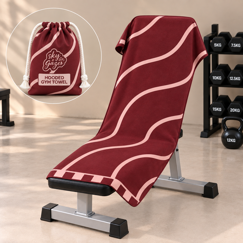 A burgundy hooded gym towel with light pink wavy stripes draped over a weight bench, with a circular inset showing the matching drawstring hoodie bag. The background shows a gym setting with dumbbells and a kettlebell.