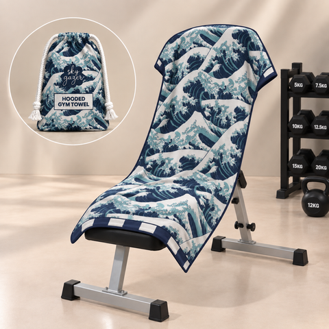 A hooded gym towel with a blue and white wave print is draped over a weight bench in a gym setting, with a circular inset showing the matching drawstring pouch.