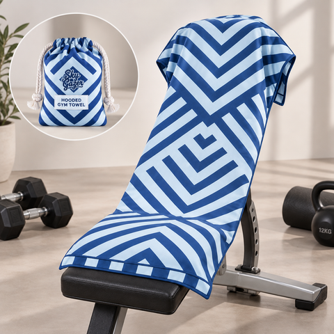 A blue and white geometric patterned hooded gym towel draped over a weight bench, with a circular inset showing the matching drawstring pouch. The scene is set in a gym with dumbbells and a kettlebell on a light wood floor.