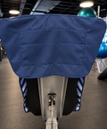 A close-up rear view of a gym exercise machine with a blue padded cover on the handlebar area, set in a gym with exercise balls and equipment in the background.