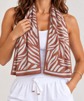 A woman wearing a terracotta brown and white patterned cooling scarf over a white tank top and white drawstring shorts, photographed against a light neutral studio background.