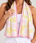 A woman wearing a pale yellow cooling towel or scarf with a pink border and large white floral print, draped around her neck over a white tank top and white drawstring shorts.