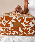 A rectangular zippered toiletry case with a brown orange and white abstract pattern, held by a person. The case features a tan leather handle, a tan leather brand patch reading 'SKY GAZER', and gold-tone hardware.