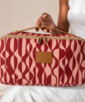 A rectangular toiletry bag with a wavy pattern in deep red and light pink, featuring a tan leather handle and a tan leather patch with the brand name 'SKY GAZER'. A person is holding the bag.