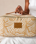 A person holds a Sky Gazer toiletry bag with a tan and white palm leaf pattern, featuring a top handle, gold zipper, and a leather brand patch.