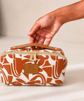 A hand holds the top handle of a rectangular zippered cosmetic bag. The bag has an abstract terracotta and white pattern, gold-tone zippers, and a tan leather patch with the brand name 'SKY GAZER'.