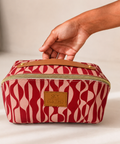 A rectangular zippered cosmetic bag with a red and pink wavy pattern, a tan leather handle, and a tan leather brand patch reading 'SKY GAZER'. A hand is holding the top handle of the bag, which is set against a neutral light beige background.
