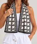 A woman wearing a black and white interlocking chain pattern cooling towel scarf draped around her neck over a white tank top and white drawstring pants, against a light gray studio background.