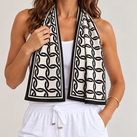 A woman wearing a black and white interlocking chain pattern cooling towel scarf draped around her neck over a white tank top and white drawstring pants, against a light gray studio background.