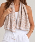 A woman wearing a white ribbed tank top and white drawstring shorts, with a beige, sage green and white patterned cooling towel draped around her neck like a scarf.