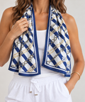 A woman models a white cooling scarf with a navy blue and gold chain-link pattern, draped around her neck over a white tank top and white drawstring pants.