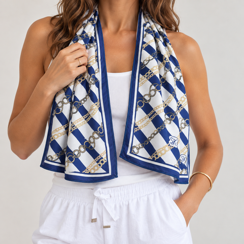 A woman models a white cooling scarf with a navy blue and gold chain-link pattern, draped around her neck over a white tank top and white drawstring pants.