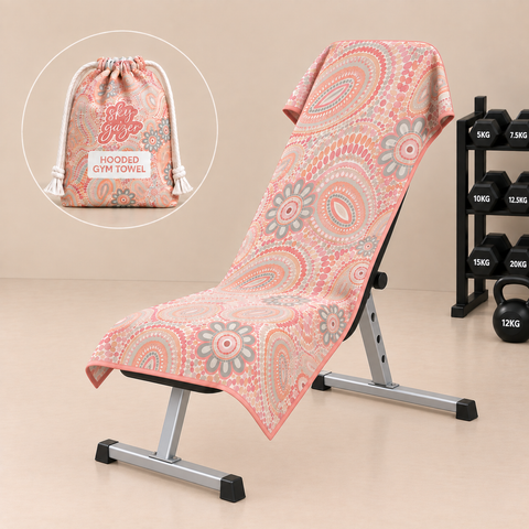 A hooded gym towel with a pink, orange, and gray paisley pattern draped over a metal bench, with a circular inset showing the matching drawstring pouch. The background is a clean beige studio setting with dumbbells on a rack.