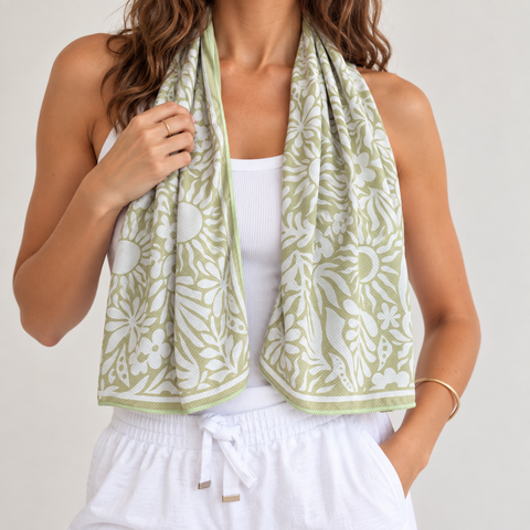 A woman models a light sage green and white patterned cotton cooling towel scarf draped around her neck, paired with a white tank top and white drawstring pants against a light gray studio background.