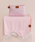 A large pink and white striped beach towel with a scalloped fringe edge, displayed on a stand, with a matching pink striped neoprene bag on top.