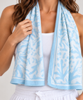 A woman wearing a white ribbed tank top and white drawstring sweatpants, with a light blue and white patterned cooling towel draped around her neck like a scarf, against a light gray studio background.