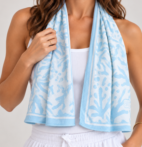A woman wearing a white ribbed tank top and white drawstring sweatpants, with a light blue and white patterned cooling towel draped around her neck like a scarf, against a light gray studio background.