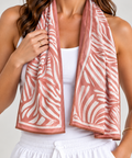 A woman wearing a brown and white patterned cooling towel scarf draped around her neck, paired with a white tank top and white drawstring shorts, against a light gray studio background.