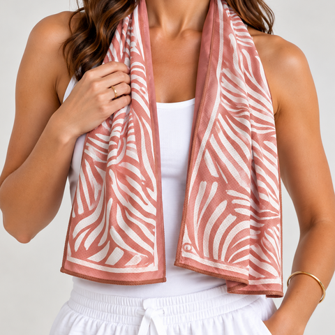A woman wearing a brown and white patterned cooling towel scarf draped around her neck, paired with a white tank top and white drawstring shorts, against a light gray studio background.